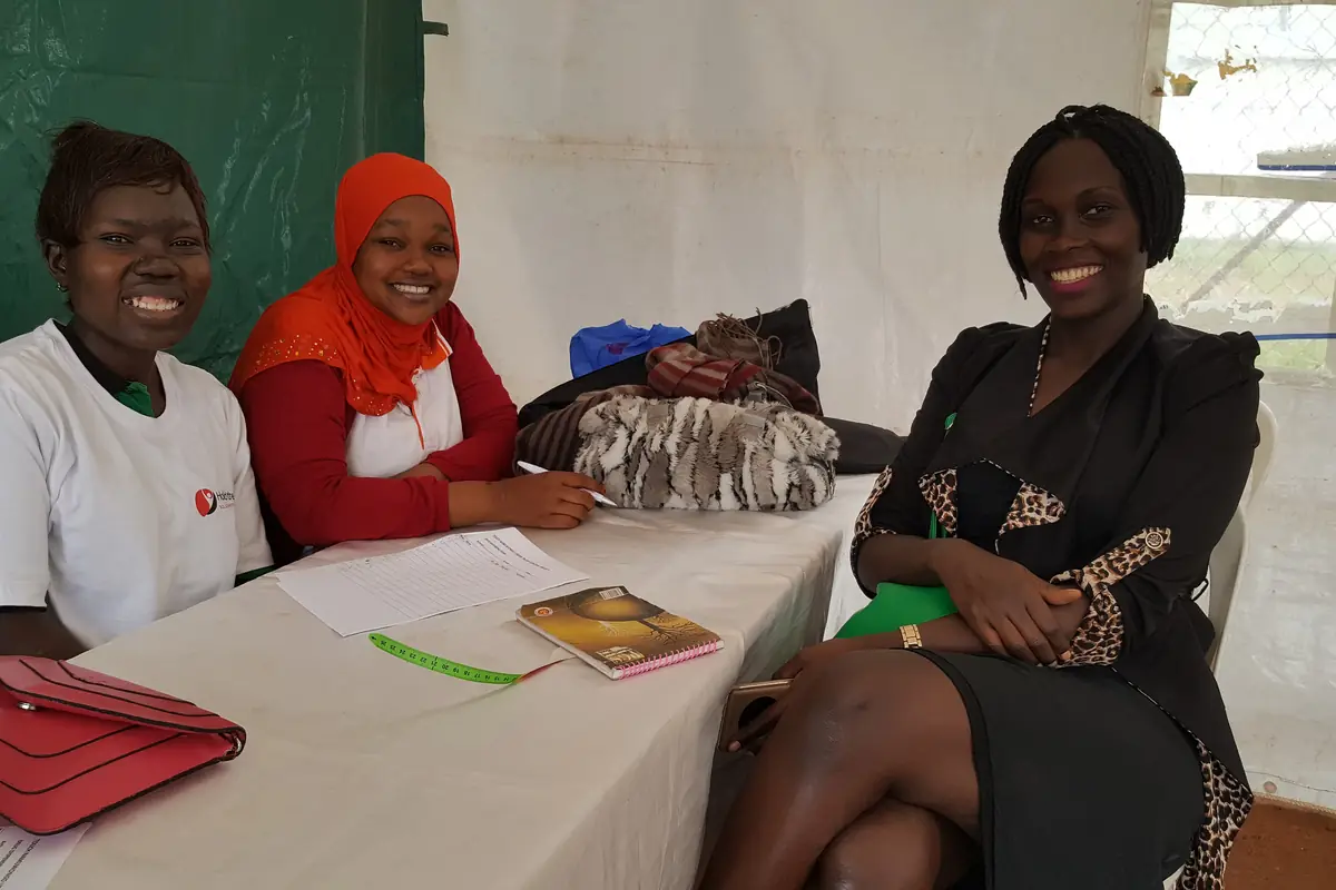 Three women smiling at a table with papers and a notebook at a work event in Kampala