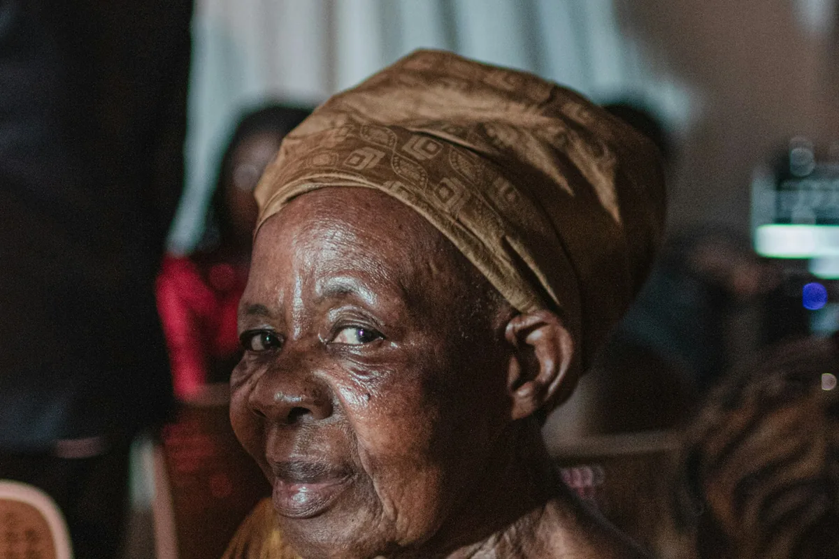 An elderly Ugandan woman in a brown headwrap smiling at a family gathering