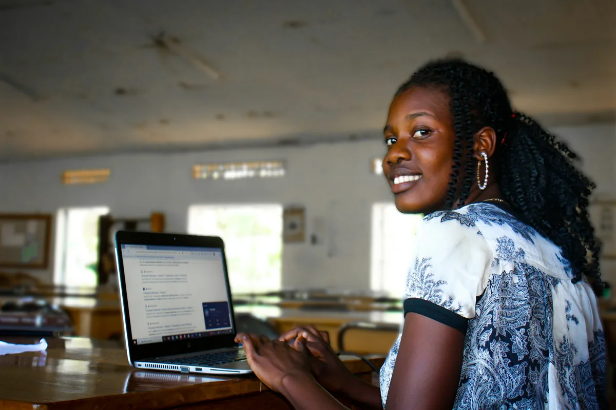 A smiling young woman typing on a laptop at a long table in a library