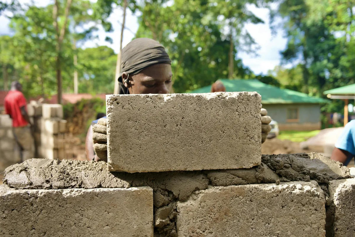 A builder placing a concrete block on a wall at an outdoor construction site in Mukono
